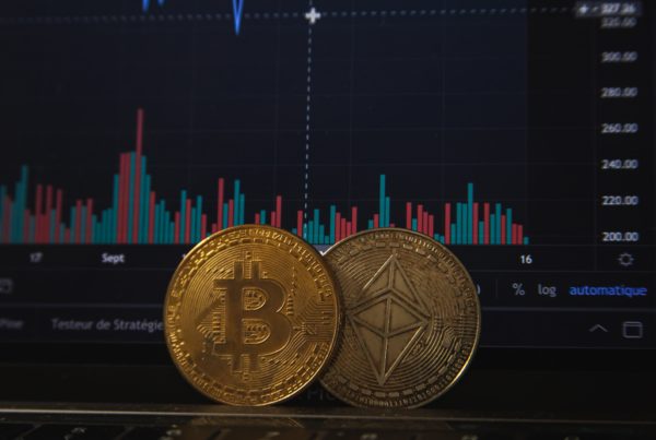 actual coins with the bitcoin logo in front of a computer screen tracking up and down graphs